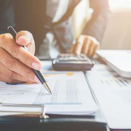 A person in business attire, representing I-Mentor Consulting, uses a pen to review financial documents and charts on a desk. A calculator and laptop sit nearby, next to eyeglasses. The well-lit scene exudes a professional atmosphere.