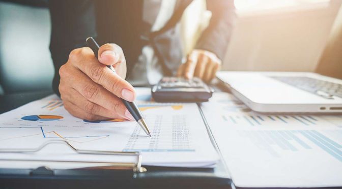 A person in business attire, representing I-Mentor Consulting, uses a pen to review financial documents and charts on a desk. A calculator and laptop sit nearby, next to eyeglasses. The well-lit scene exudes a professional atmosphere.